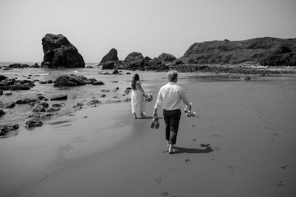 Black and white photograph of two people walking on a scenic beach with rocky outcrops.