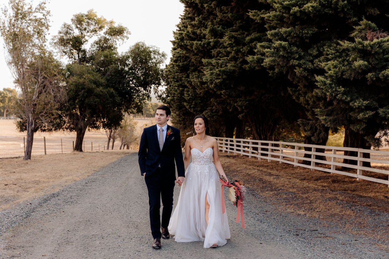 Couple in wedding attire walking together down a country dirt road lined with white fencing and trees.