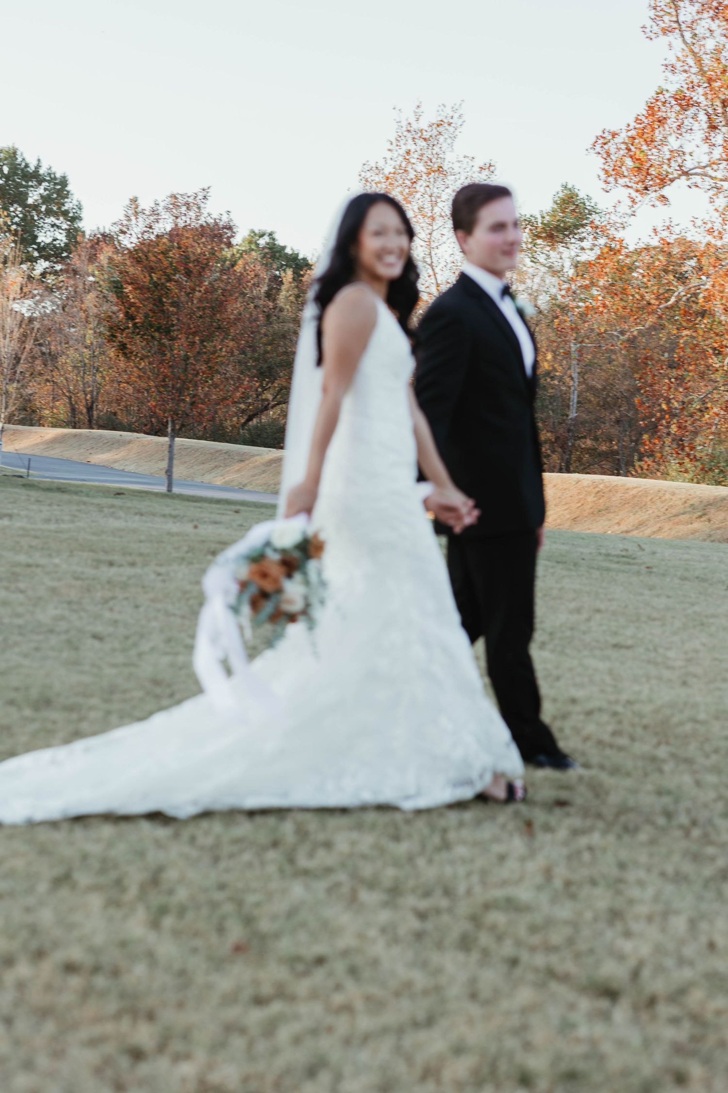 A couple in wedding attire sit together on grassy field during autumn with orange leaves in the background.