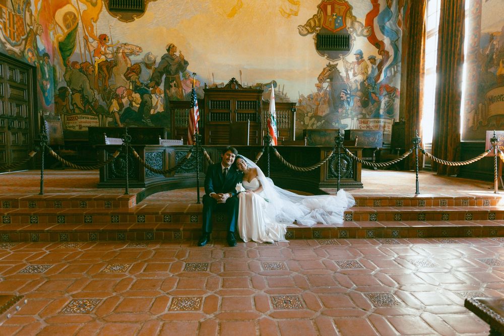 A serene couple in wedding attire rests on an antique bench inside an ornate historic building with tiled floors.