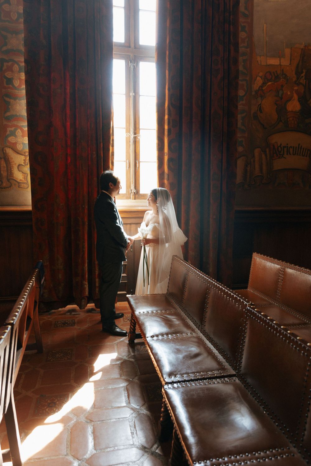 A couple shares an intimate moment during wedding by a tall window with dramatic sunlight streaming in, surrounded by vintage furniture.