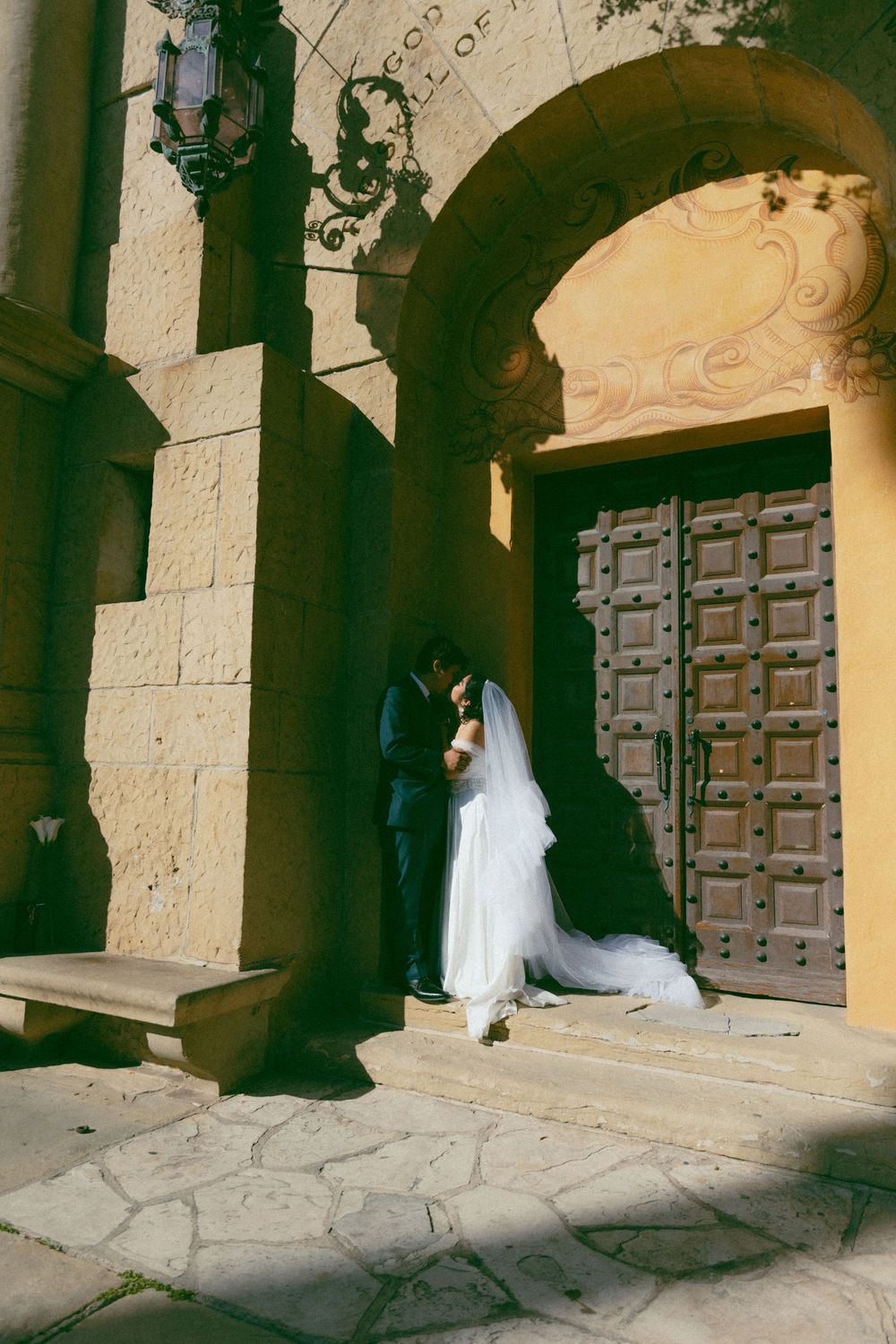 A wedding couple stands in shadow under a grand stone archway with ornate wooden doors at a traditional church entrance.