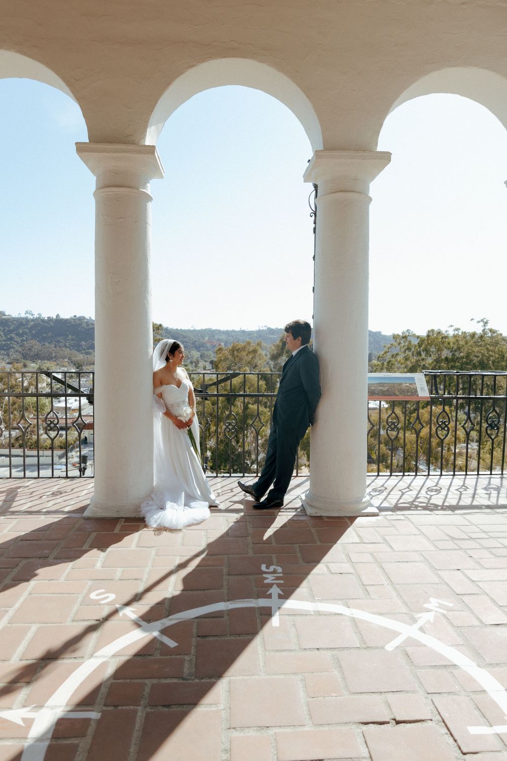 Bride and groom share a romantic first look on a sunlit balcony with white arches and scenic views at an outdoor wedding venue.