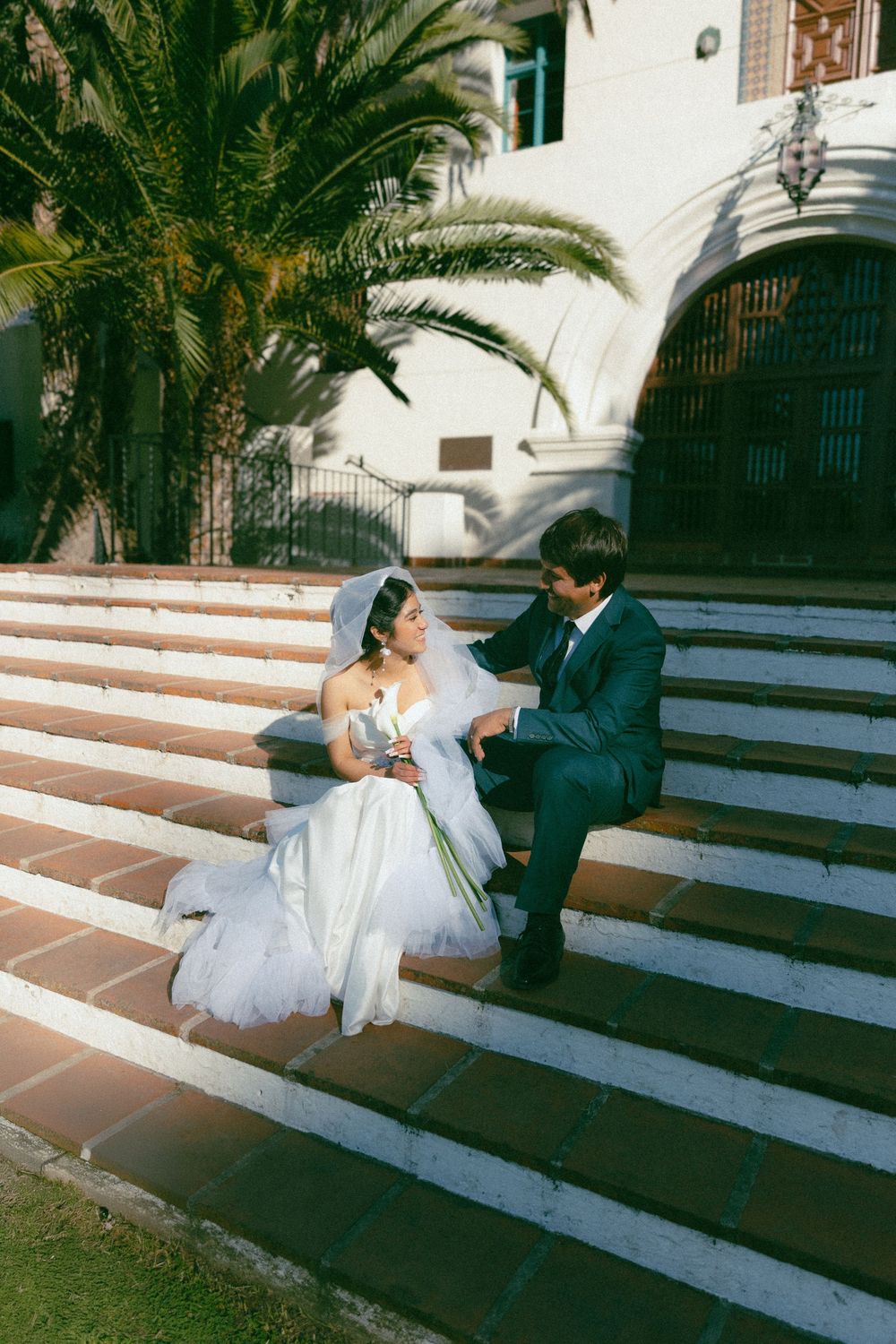 Bride and groom sitting on church steps, bride in white gown with veil, groom in navy suit, palm trees behind.