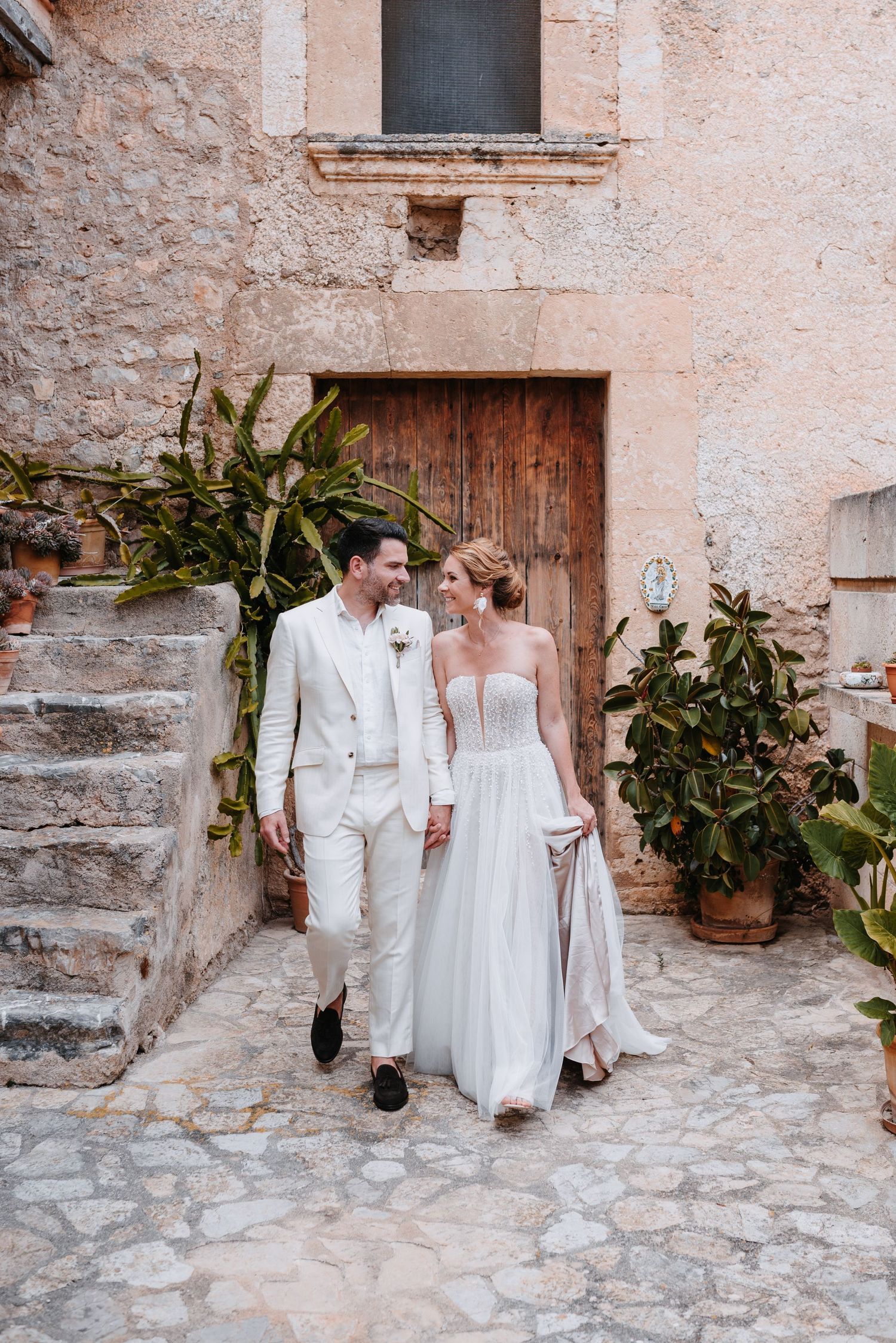 Ein Paar in eleganter weißer Hochzeitskleidung spaziert gemeinsam auf einem rustikalen Kopfsteinpflasterweg mit Pflanzen und Steinmauer.