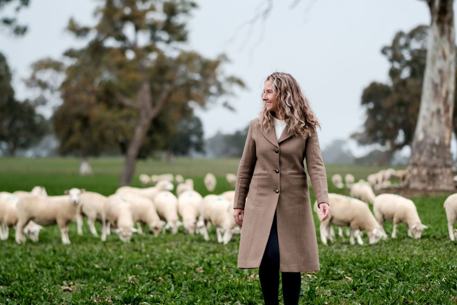 A person in brown coat standing in green field with grazing sheep.