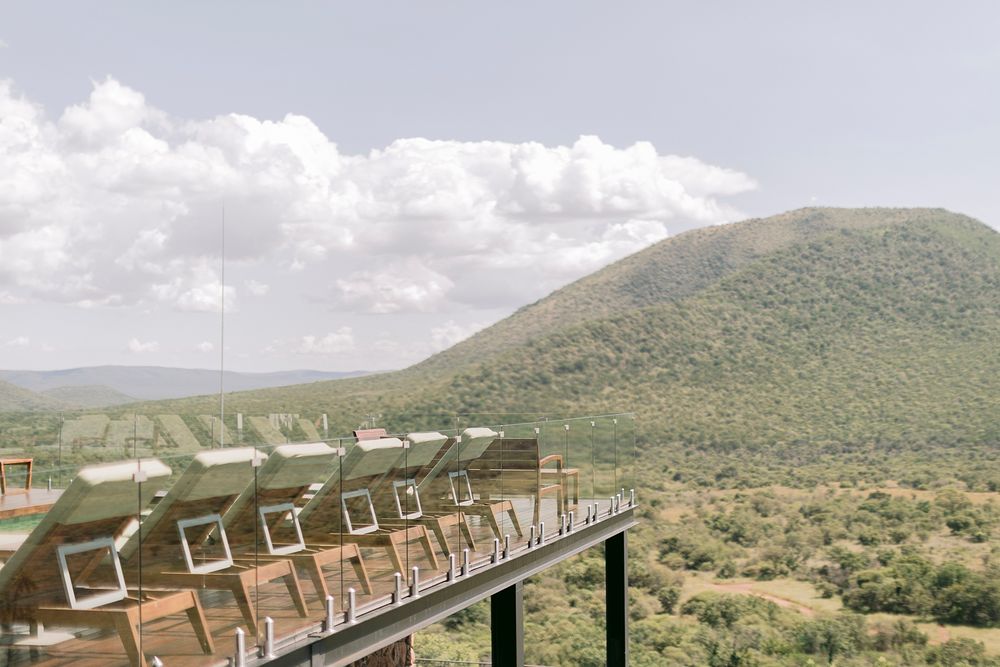 Luxury safari lodge deck chairs overlooking lush green hills and savanna landscape under cloudy sky.