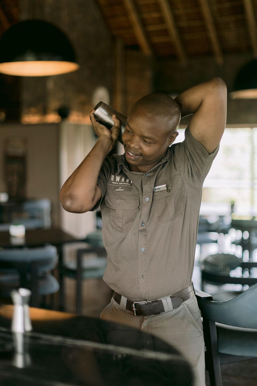 African bartender in safari uniform shaking cocktails at an upscale lodge bar interior.
