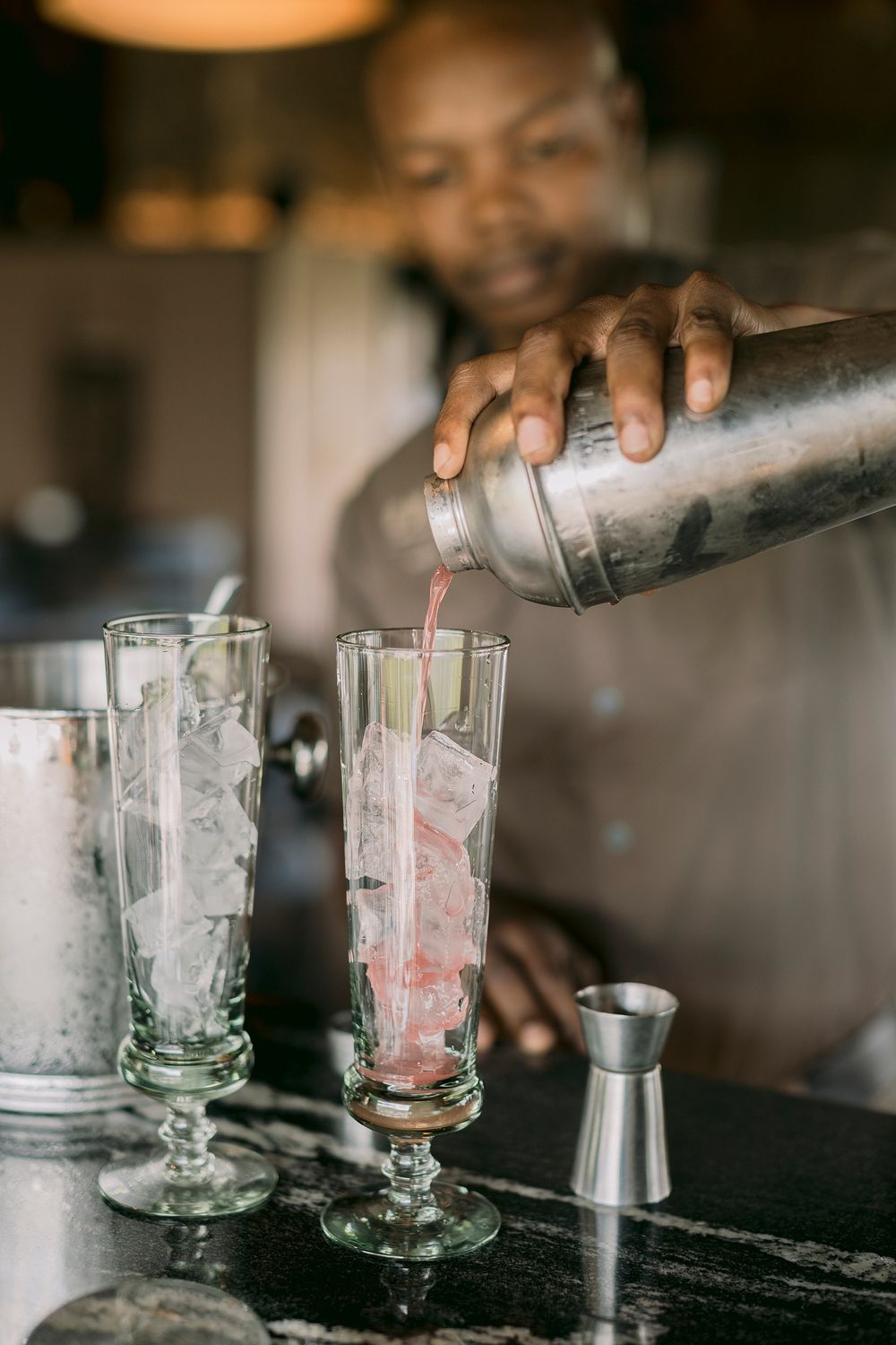 Bartender pouring pink cocktail from shaker into ice-filled glasses at a luxury safari lodge bar.