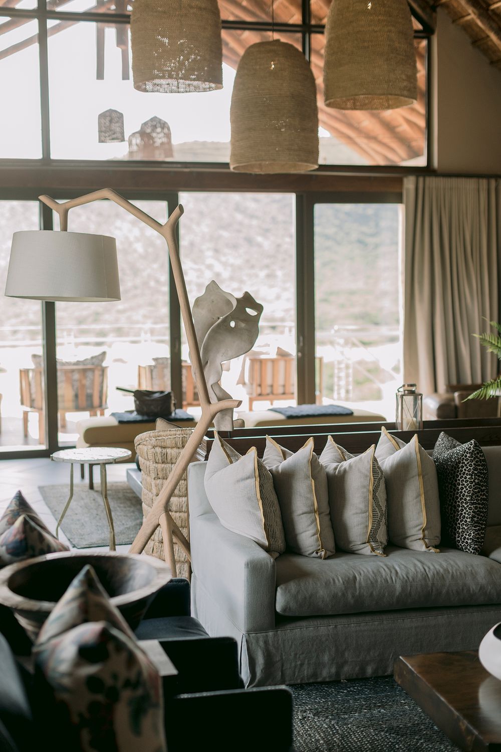 Elegant safari lodge lounge with neutral cushioned sofa, woven pendant lights and mountain views.