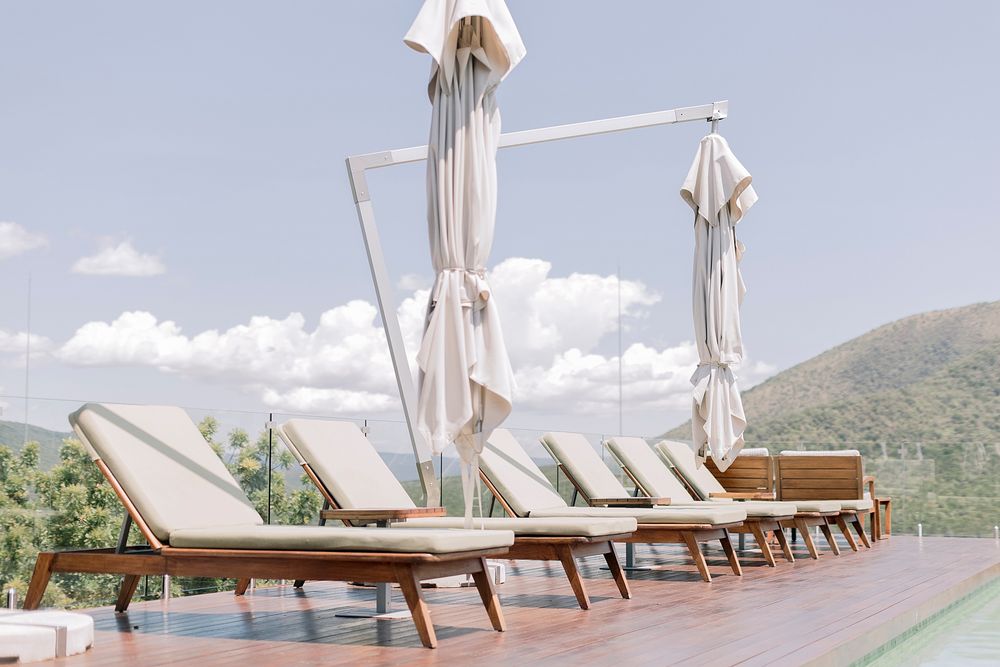 Row of wooden sun loungers with white umbrellas beside a pool overlooking mountain scenery.
