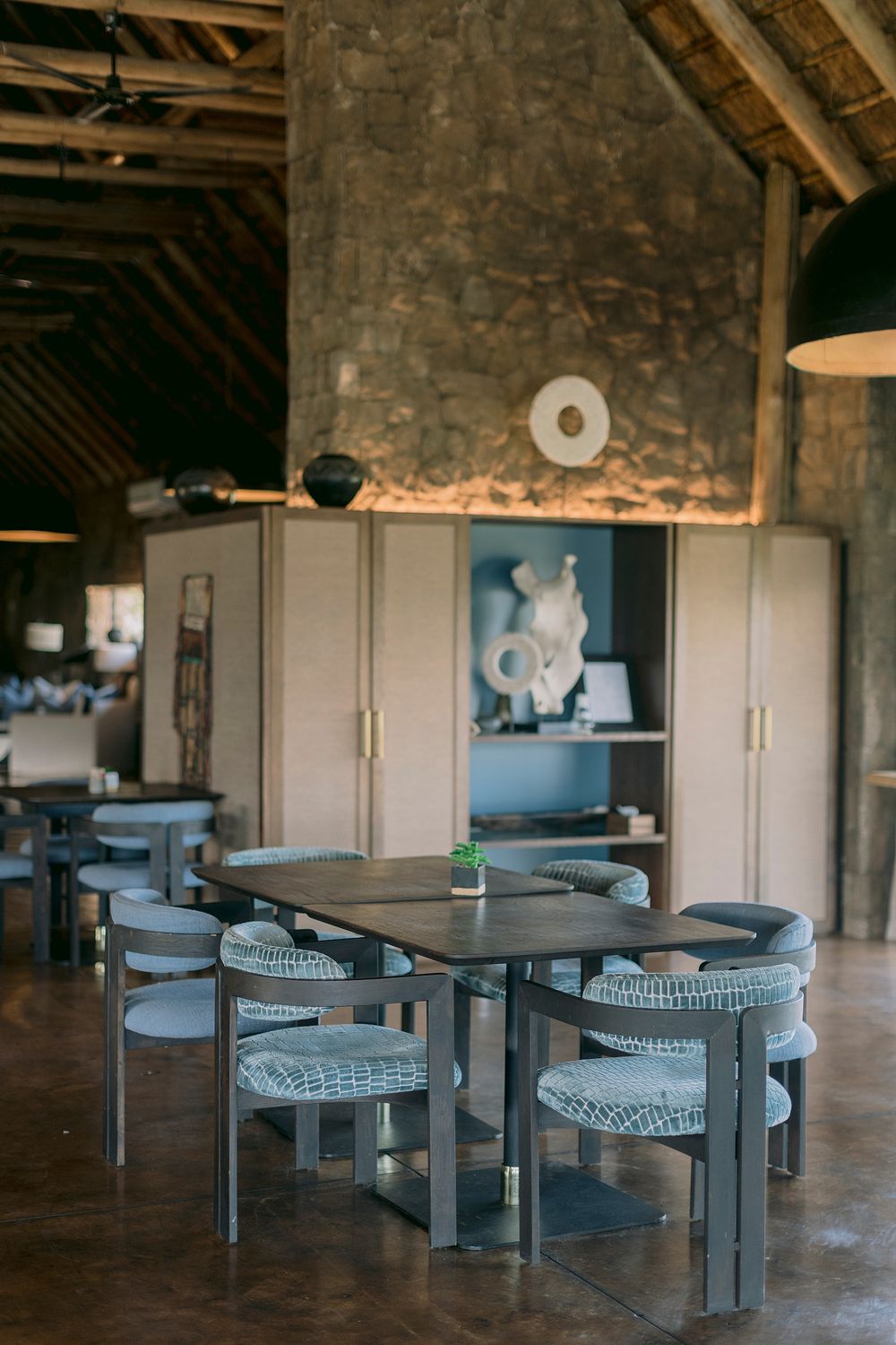Elegant safari lodge dining area with stone walls, thatched ceiling, and modern grey chairs around wooden tables.