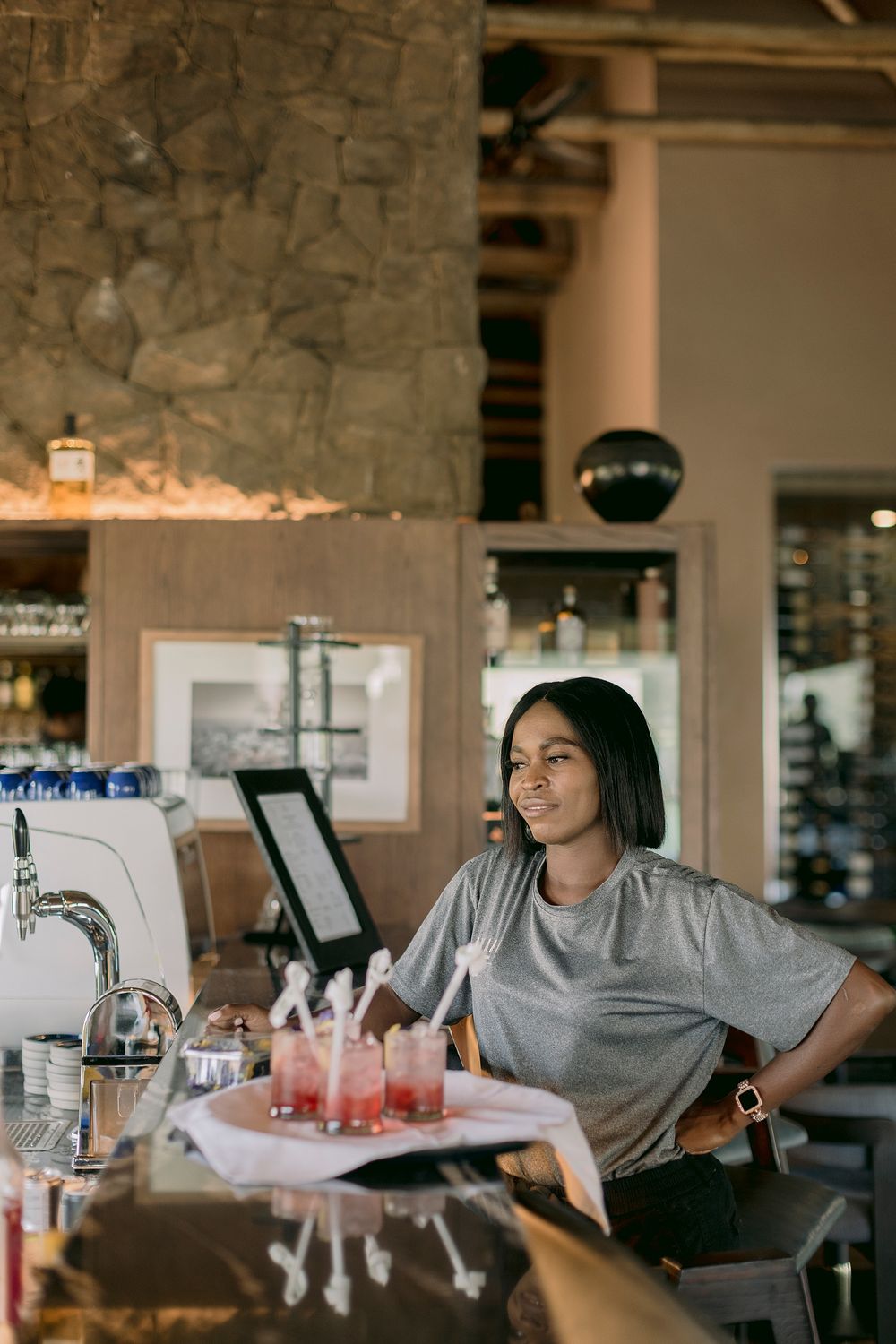 Smiling bartender standing behind a bar counter with a tray of pink cocktails garnished with flowers.