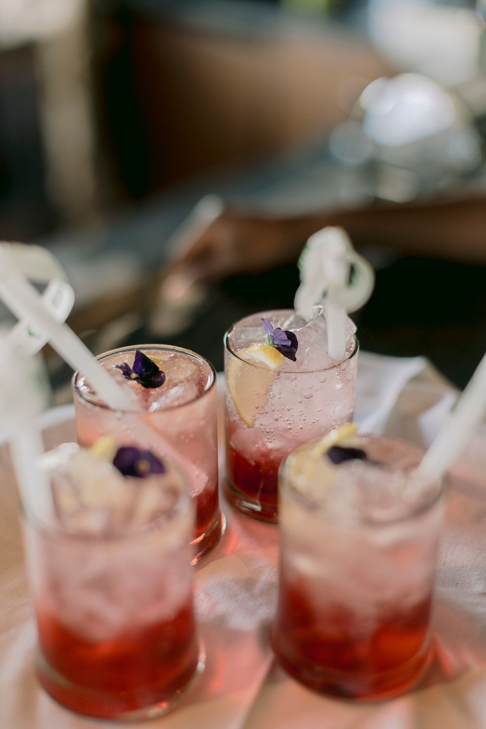 Four pink cocktails with ice, lemon slices, purple flower garnishes, and white paper straws on a tray.