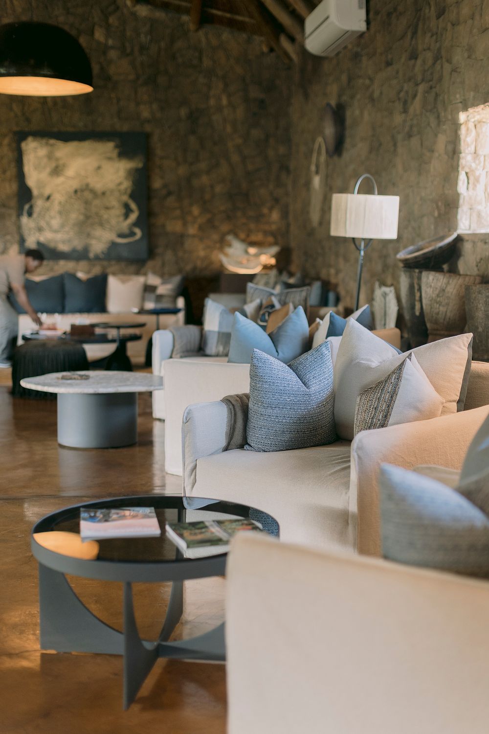 Luxurious safari lodge lounge with cream sofas, blue cushions, stone walls, and warm ambient lighting.