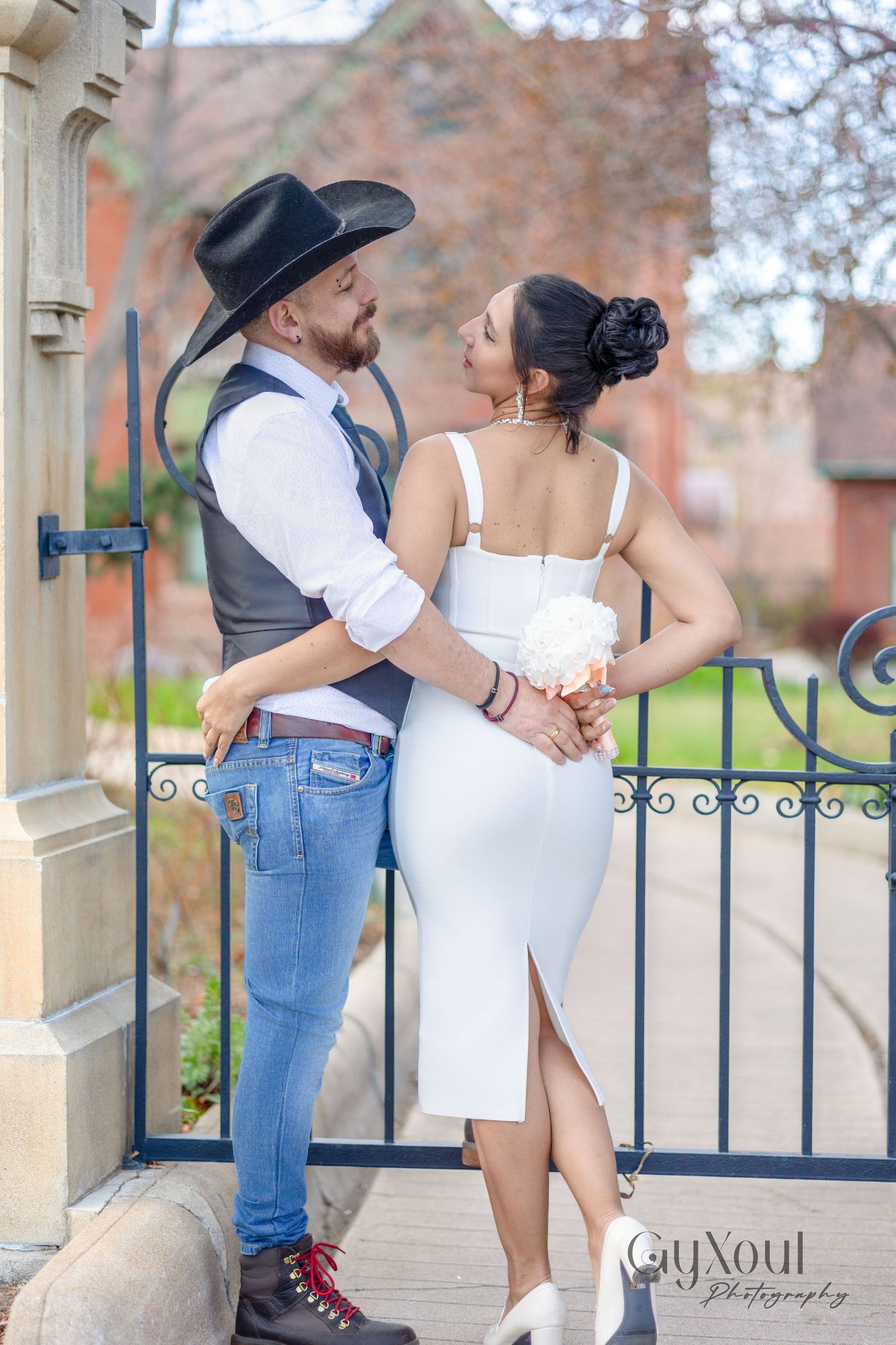 A romantic couple embraces by an iron gate, with the person wearing a black cowboy hat and vest standing with someone in white.