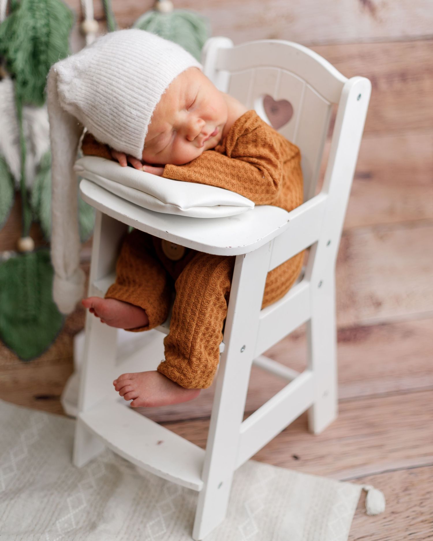 Sleeping baby wearing a knit beanie and brown sweater rests peacefully in a white wooden high chair.