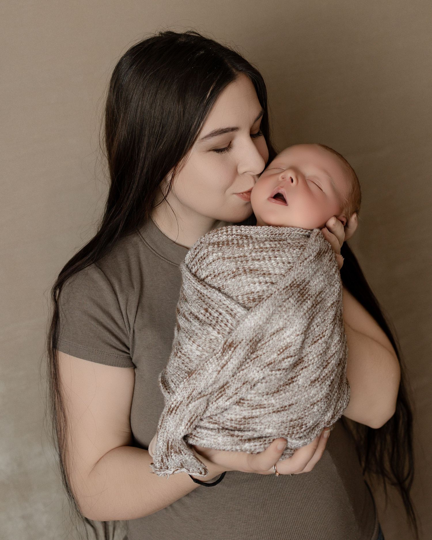 A tender moment captured as a parent gently kisses a sleeping newborn baby wrapped in a knitted beige blanket.