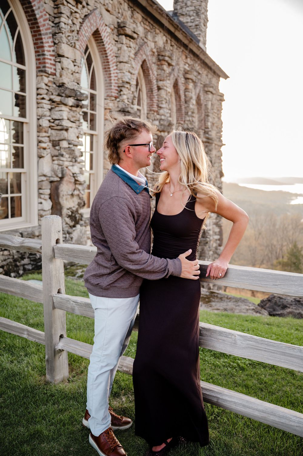 Couple smiling and embracing near a rustic wooden fence with a stone building backdrop at golden hour.