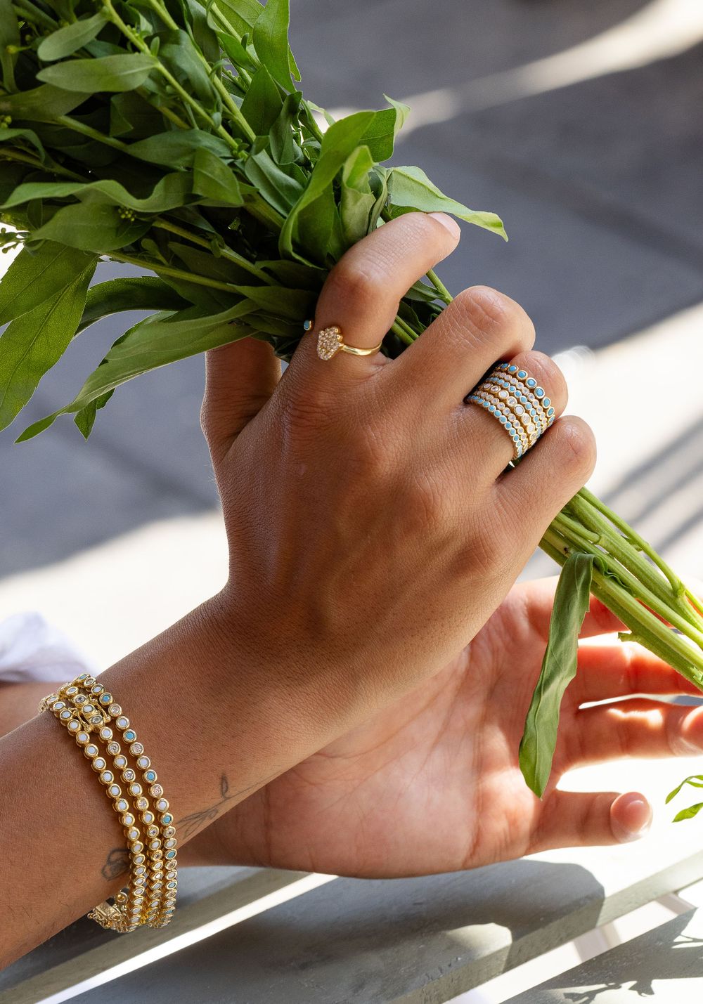 Hand adorned with elegant rings and pearl bracelet holding fresh green herbs in natural sunlight.