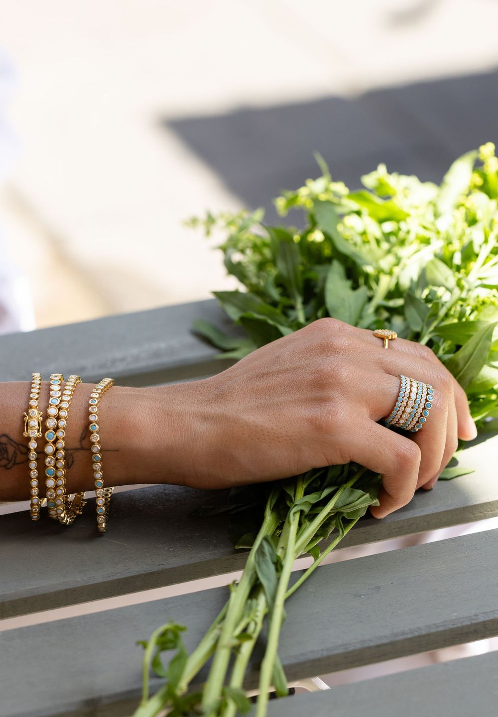 Hand wearing gold bracelets and rings holds fresh green herbs against a dark gray background.
