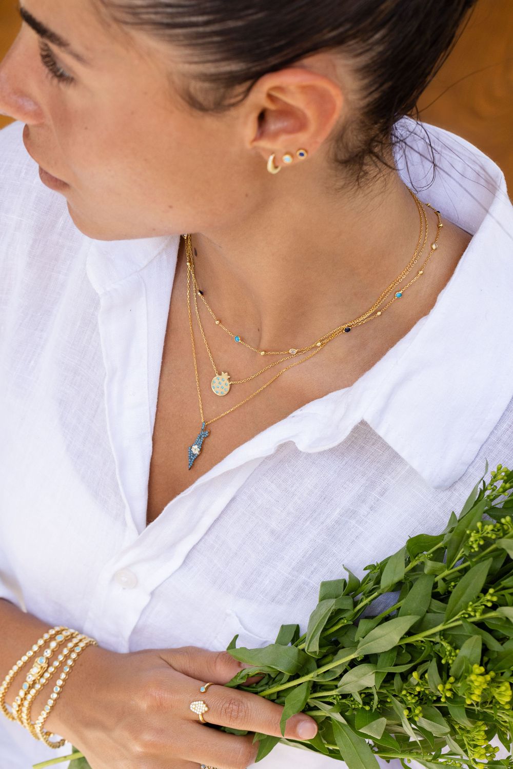 Close-up of layered gold necklaces with pendants worn with white linen shirt and pearl bracelet accessories.