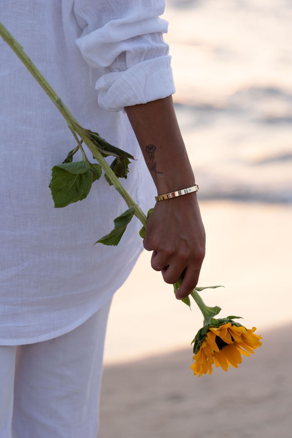 A hand wearing a white outfit holds a vibrant yellow sunflower with a simple beaded bracelet against a soft background.