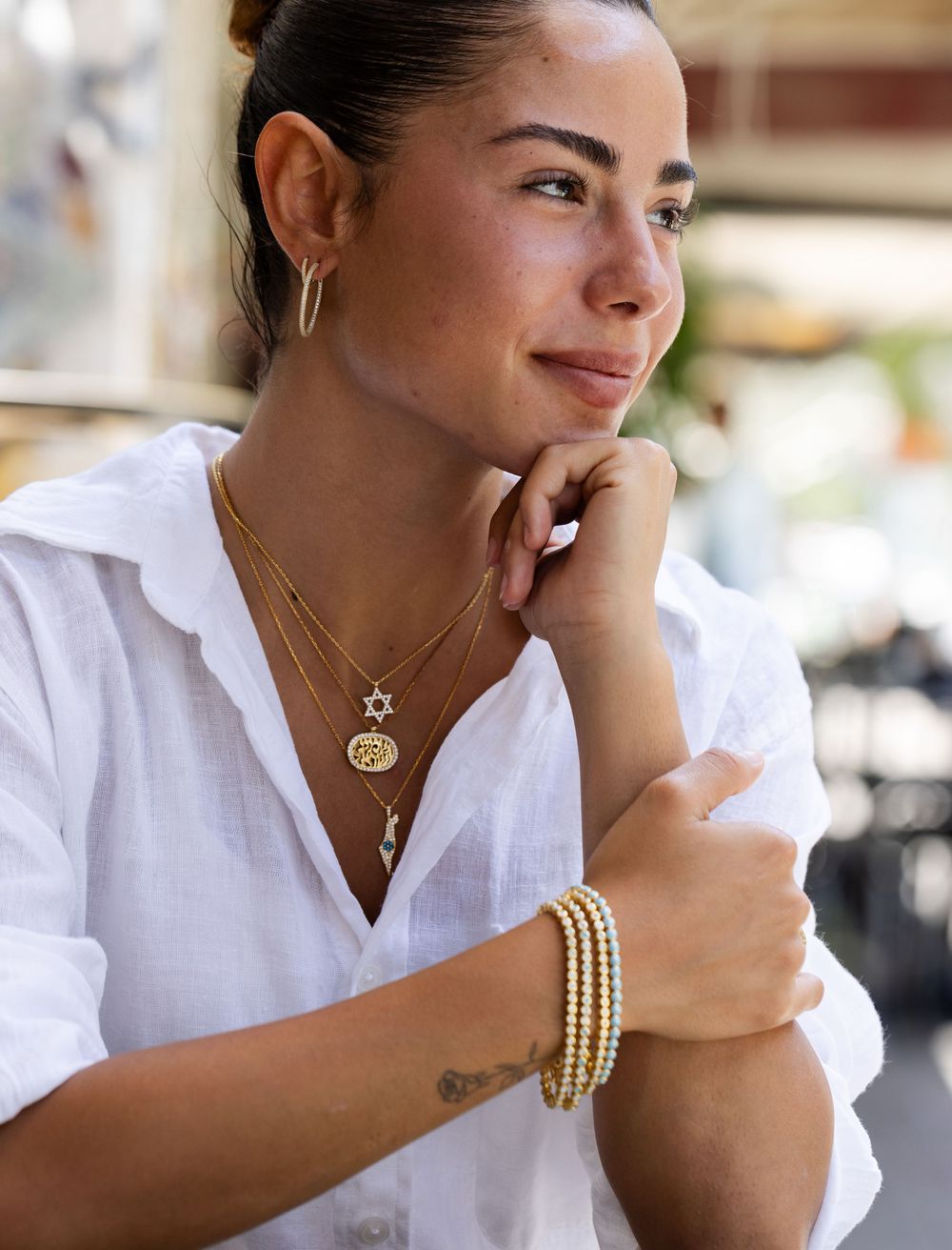 Woman in crisp white button-down shirt smiling while wearing layered gold necklaces and pearl bracelets in outdoor setting.