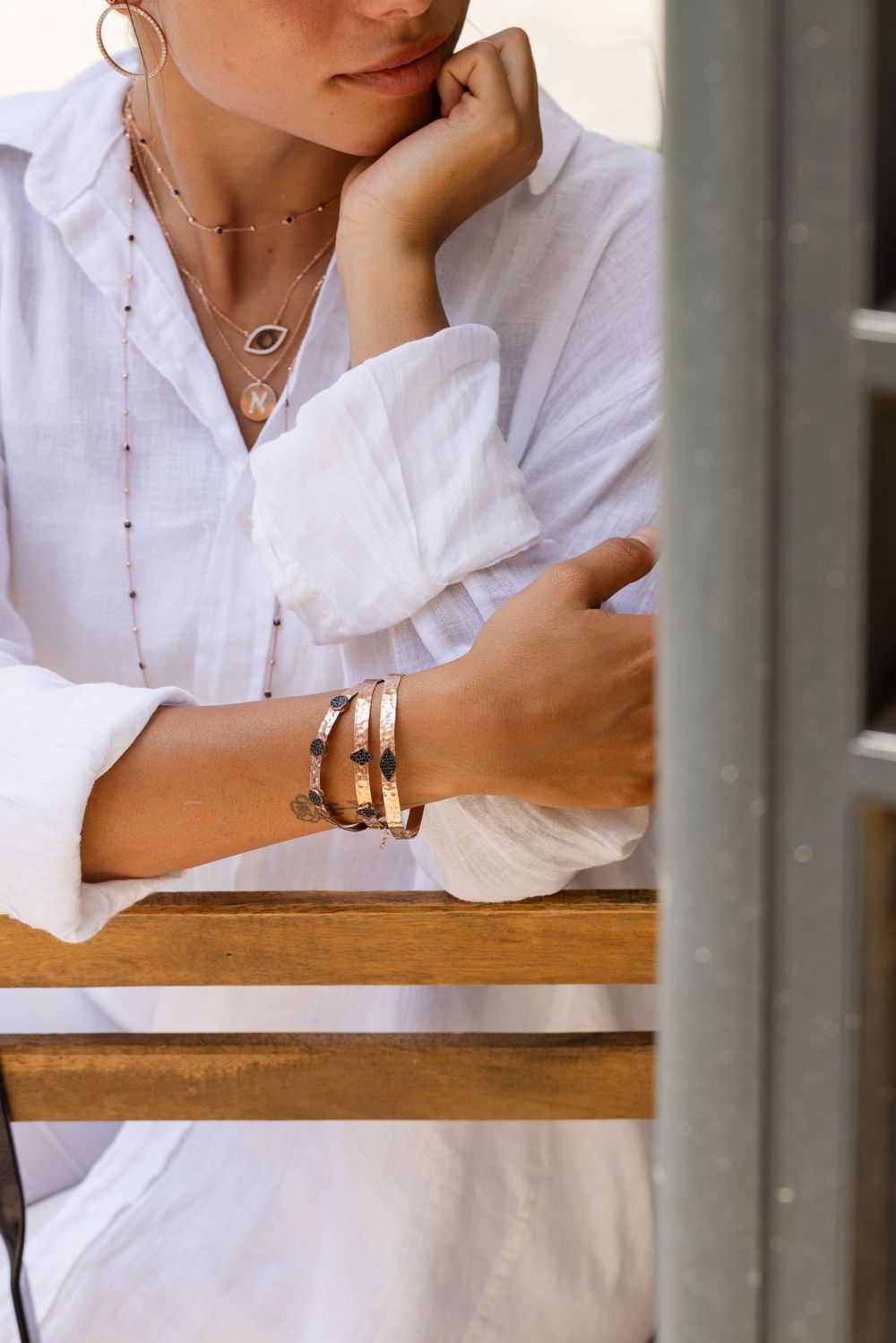 Elegant layered necklaces and bangles accessorize a crisp white button-down shirt while leaning on wooden surface.