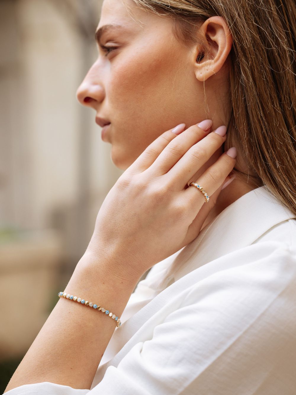 Elegant hand adorned with dainty rings and bracelet touching neck against neutral background.
