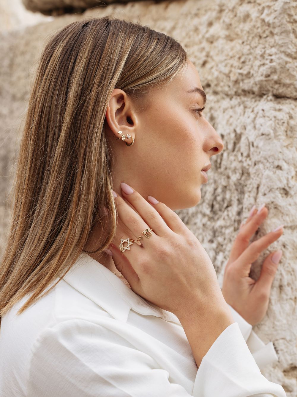 Profile view showing long straight hair and delicate jewelry against a textured stone wall background.