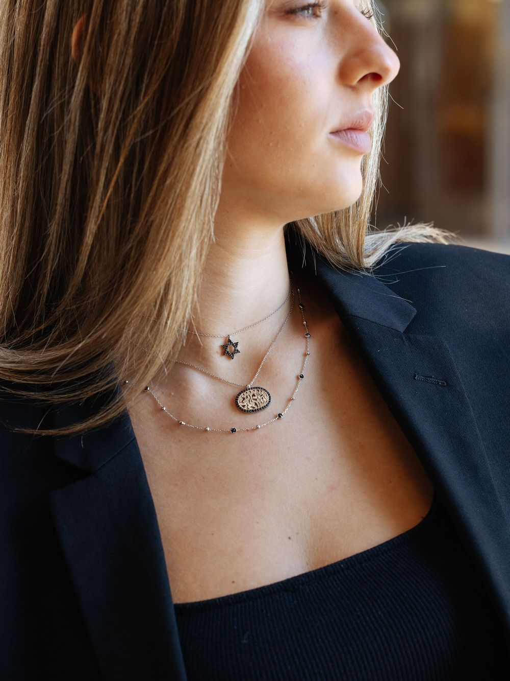 Woman in black blazer wearing delicate layered necklaces with pendant charms against a blurred background.