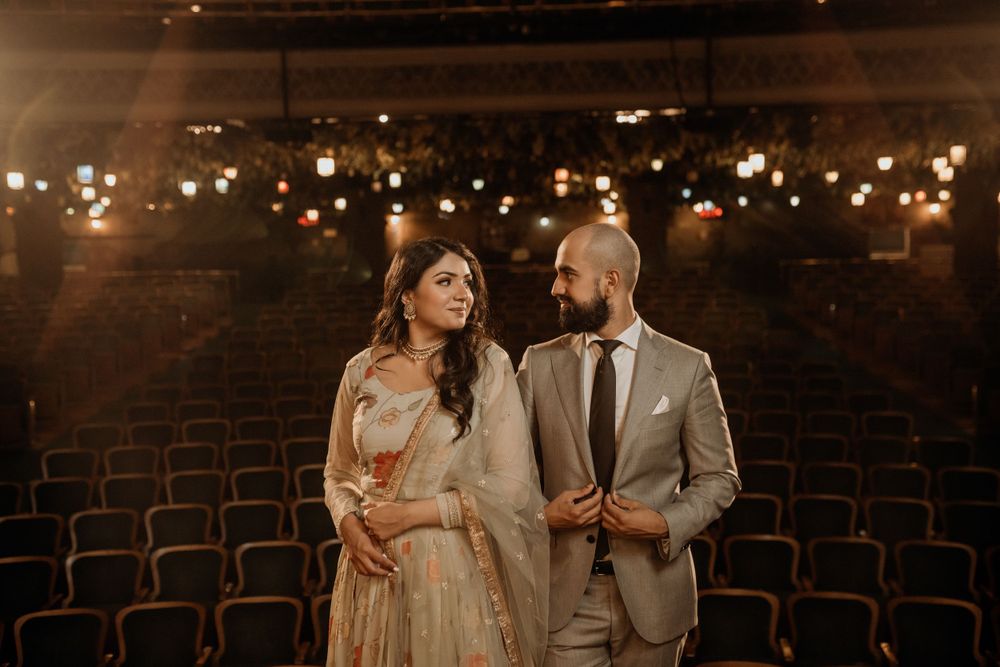 A couple in traditional wedding attire stands together in an empty vintage theater with warm glowing lights in the background.
