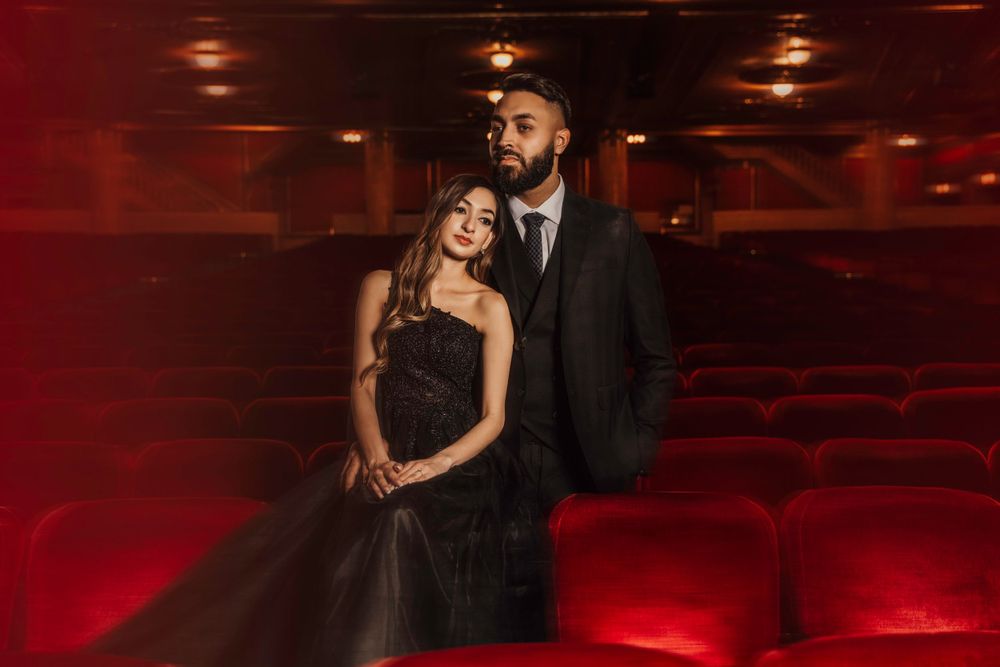 A couple in formal attire poses romantically in an empty theater with dramatic red lighting and plush seats.
