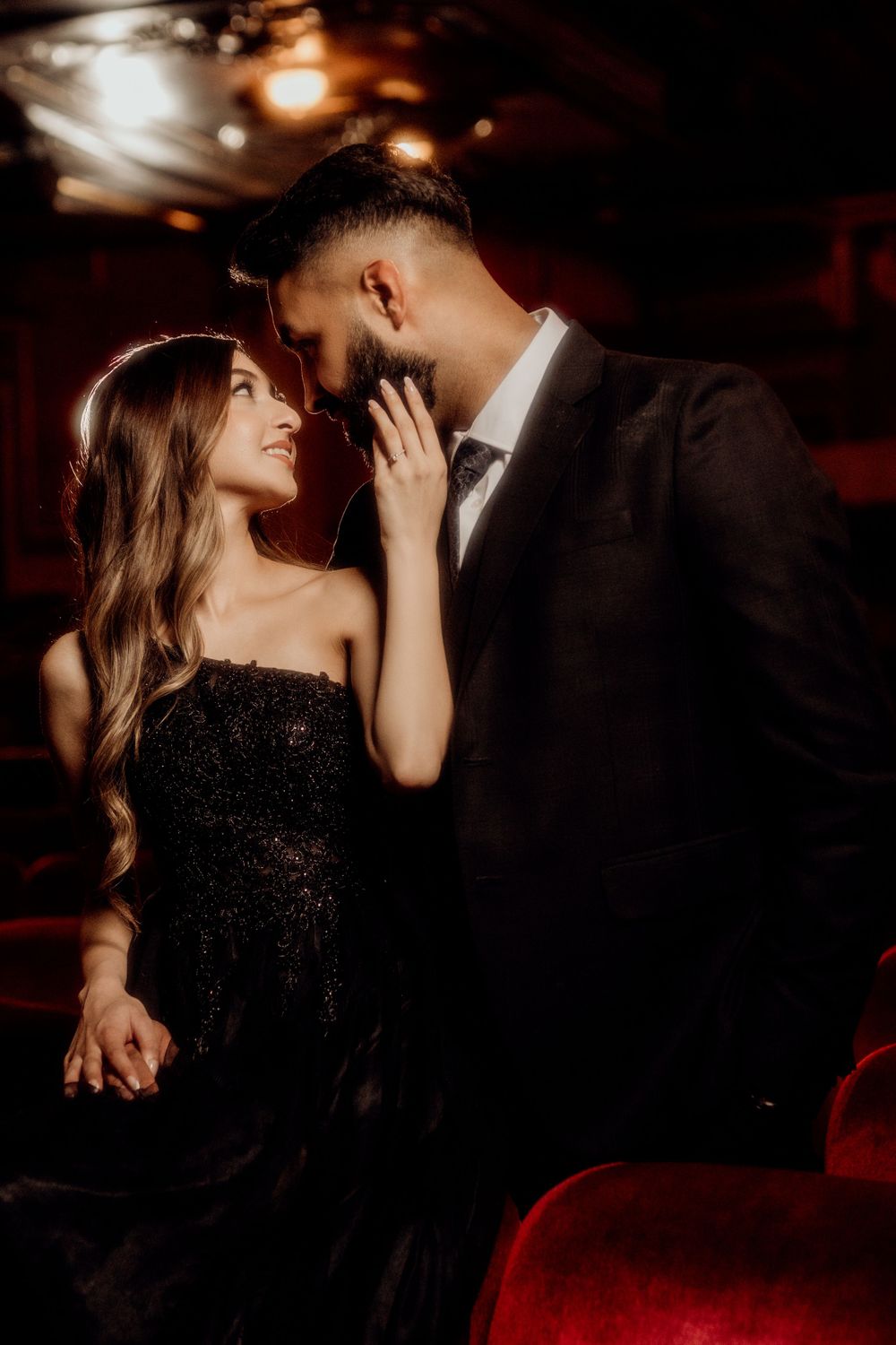Romantic couple in formal attire share an intimate moment in dark moody lighting at an elegant venue.