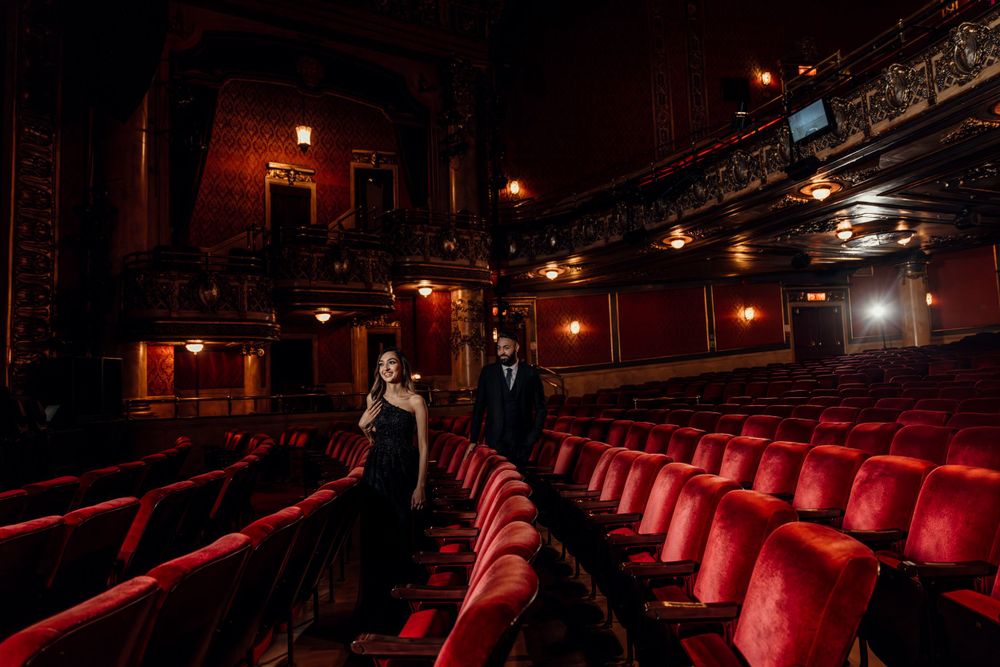 A romantic couple stands in the aisle of a dimly lit vintage theater with red velvet seats and ornate architectural details.