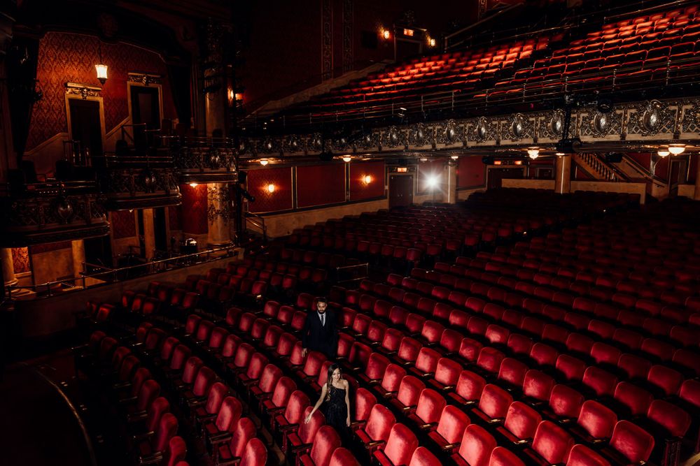 Empty red velvet seats in a dark theater auditorium with ornate decor and ambient lighting creating a dramatic atmosphere.