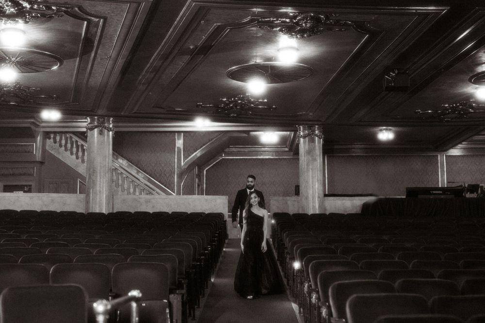 A couple stands in silhouette in an empty vintage theater with ornate ceiling details and rows of empty seats in black and white.