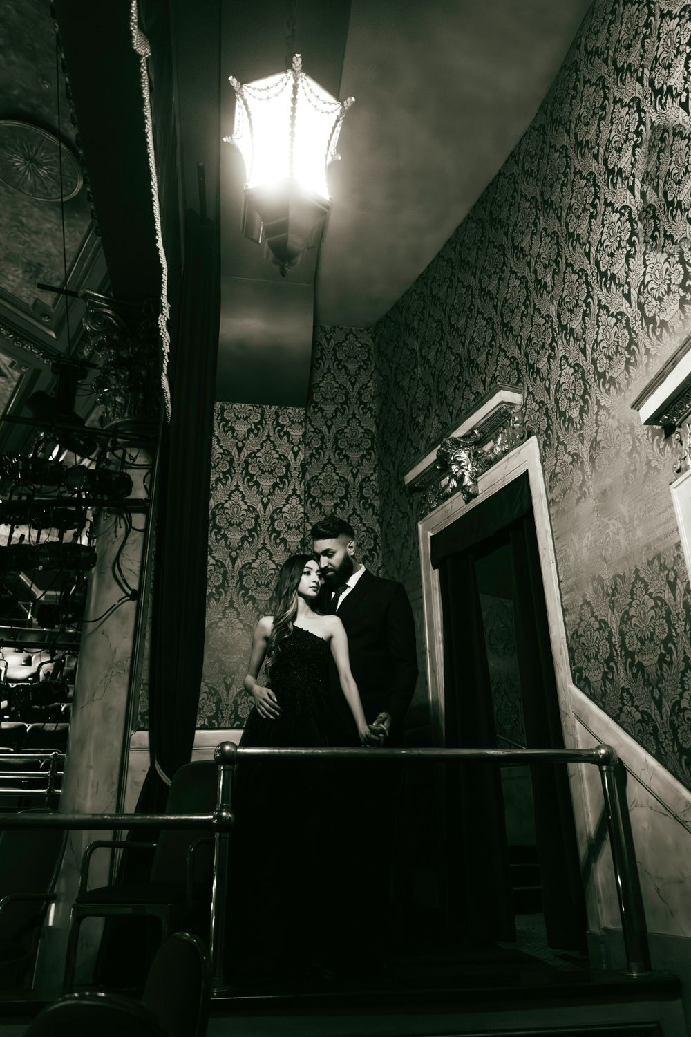 A couple embraces on a staircase in a dimly lit vintage interior with ornate wallpaper and an antique lamp above.