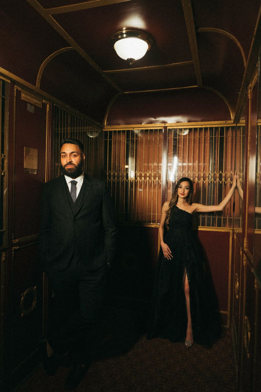 A glamorous couple in evening wear poses dramatically in a vintage elevator with gold details and warm lighting.