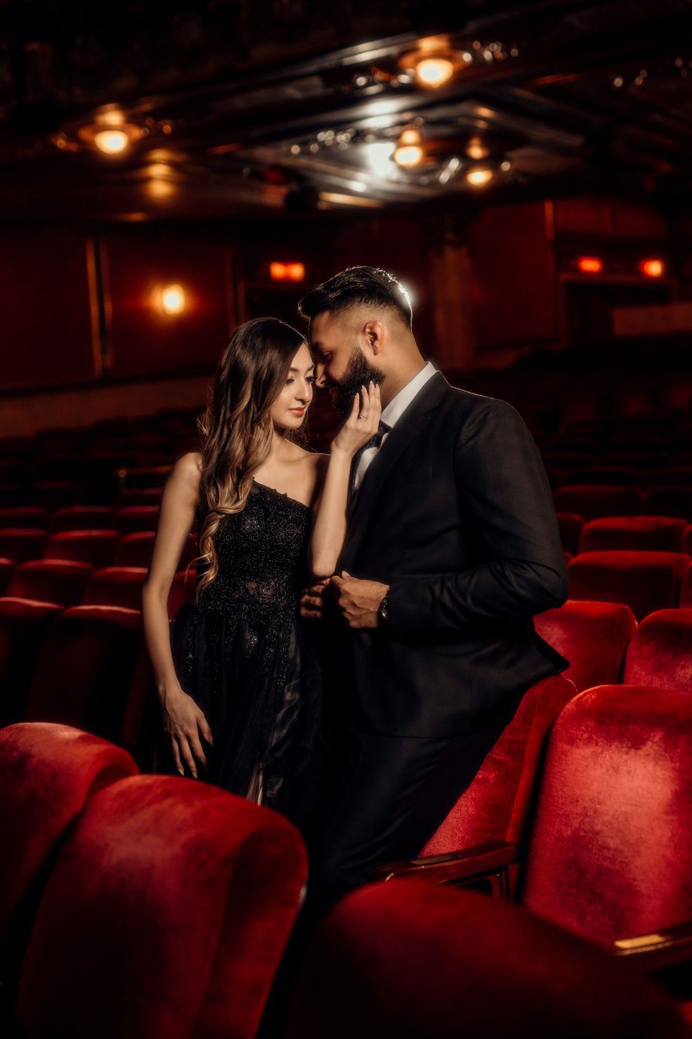 Romantic couple in formal attire sharing an intimate moment between red velvet theater seats in moody lighting.