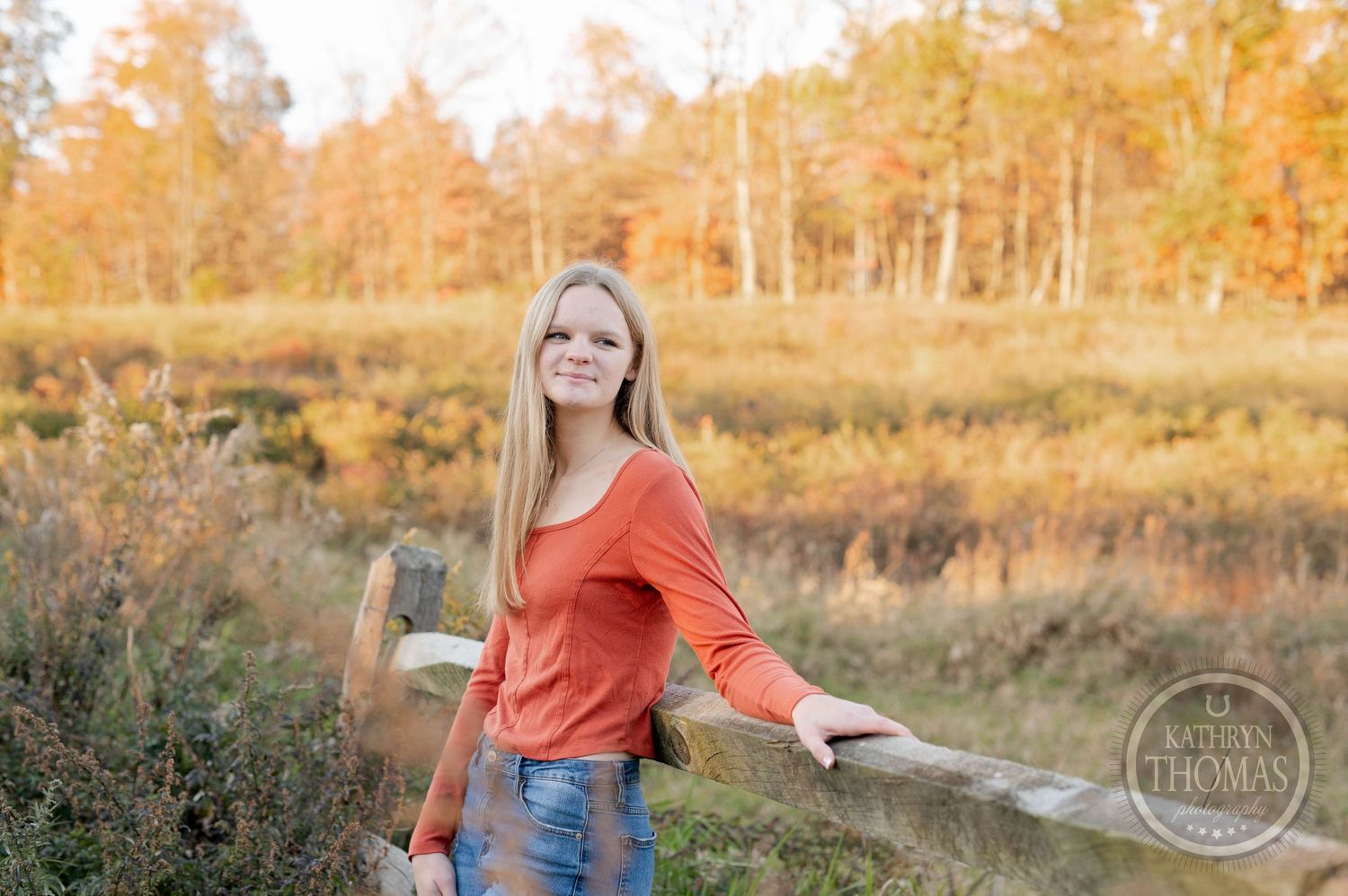 Someone in orange sweater poses by rustic fence with fall foliage creating natural backdrop.