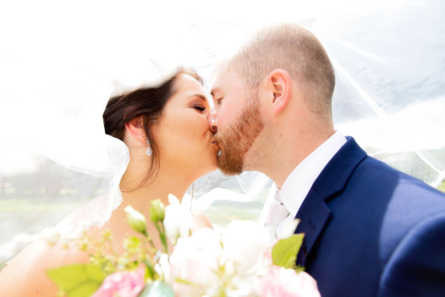 A tender moment between newlyweds sharing their first kiss as a married couple against a bright outdoor backdrop.