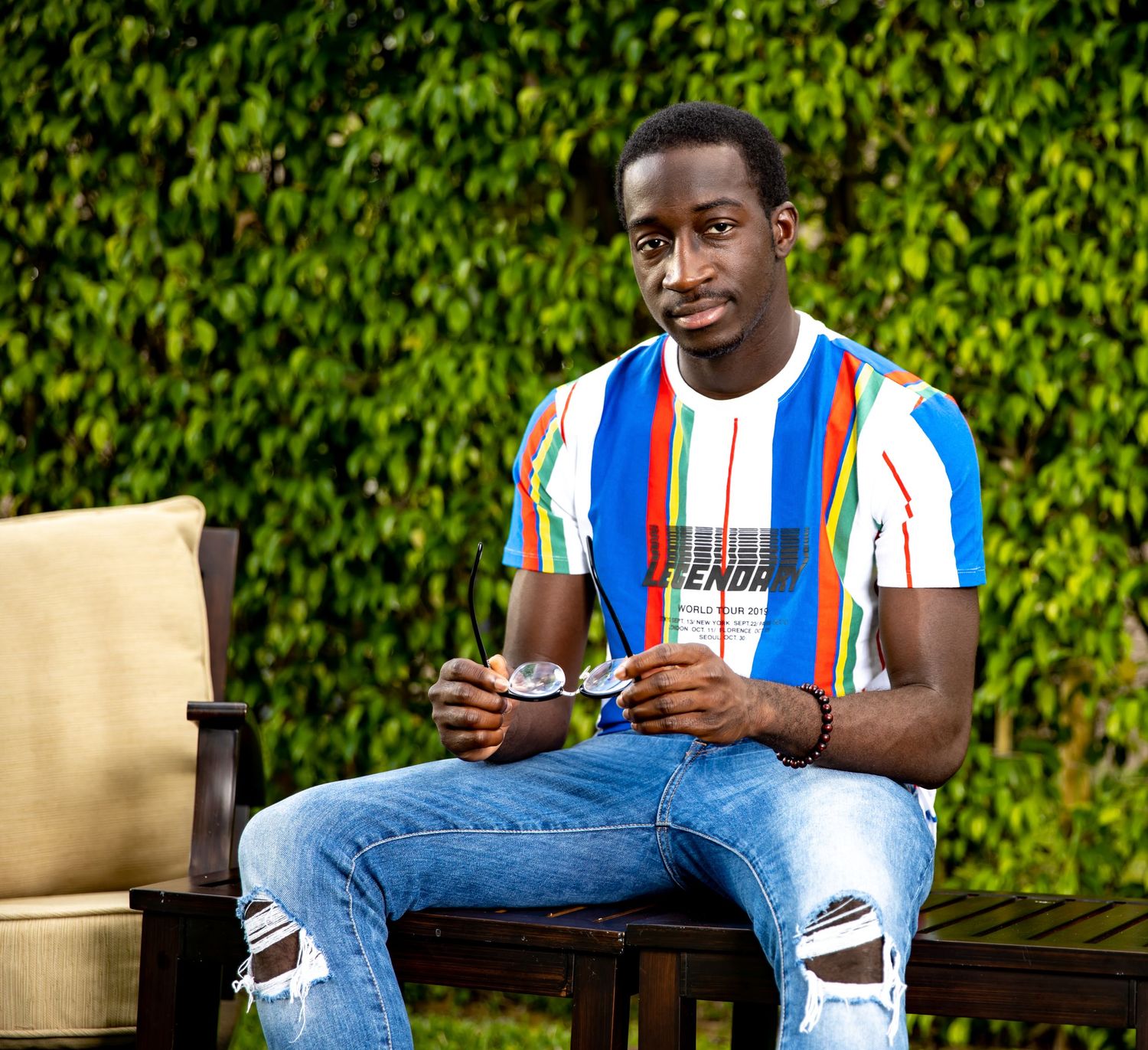 Person in striped shirt and ripped jeans sits casually in outdoor garden setting.