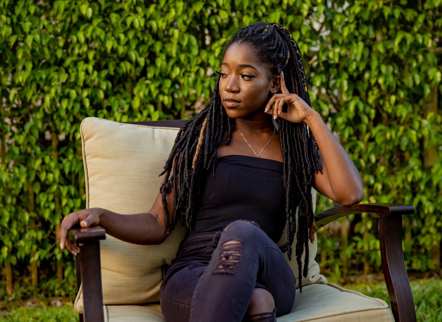 A woman with long braids relaxes outdoors in a patio chair against a lush green hedge backdrop.