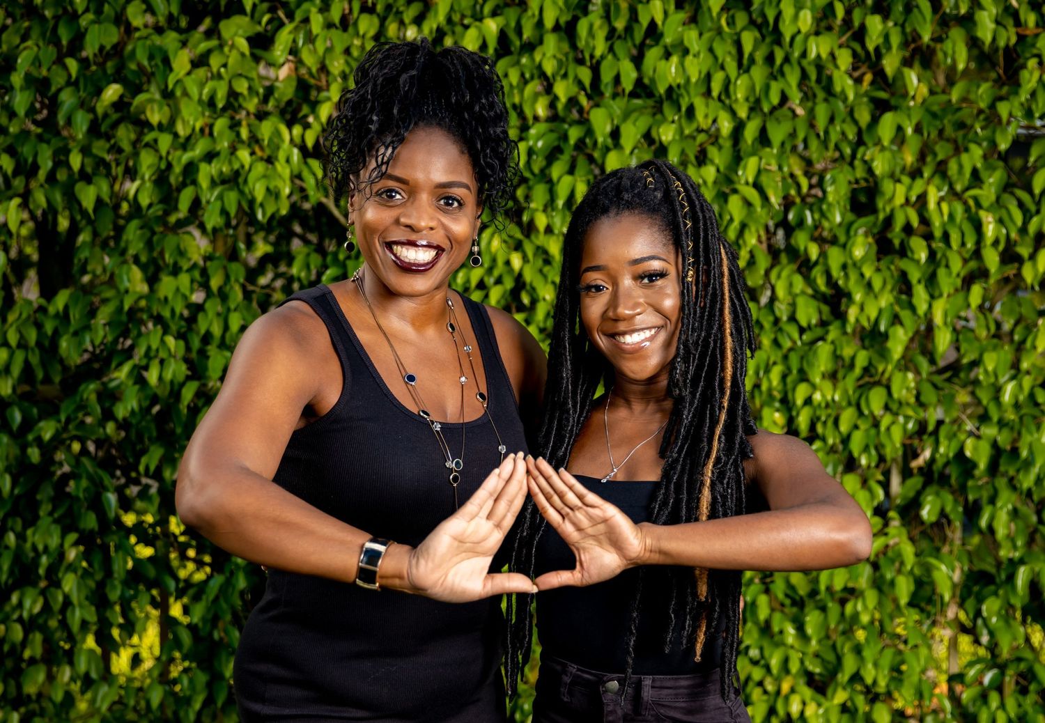 Two people in black tops smile while forming a heart shape with their hands against a lush green ivy background.