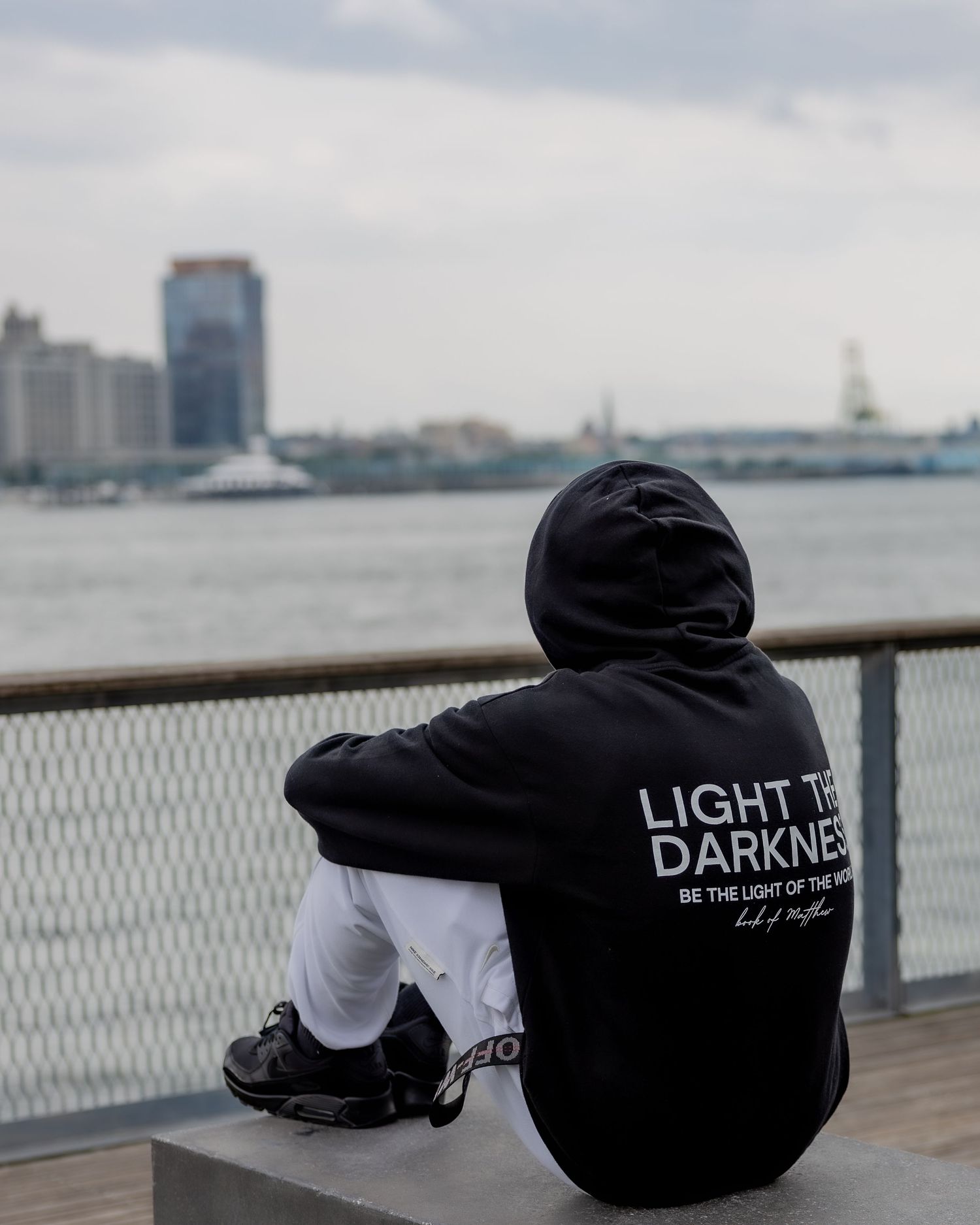 Person in black hoodie sitting alone on pier overlooking city harbor.