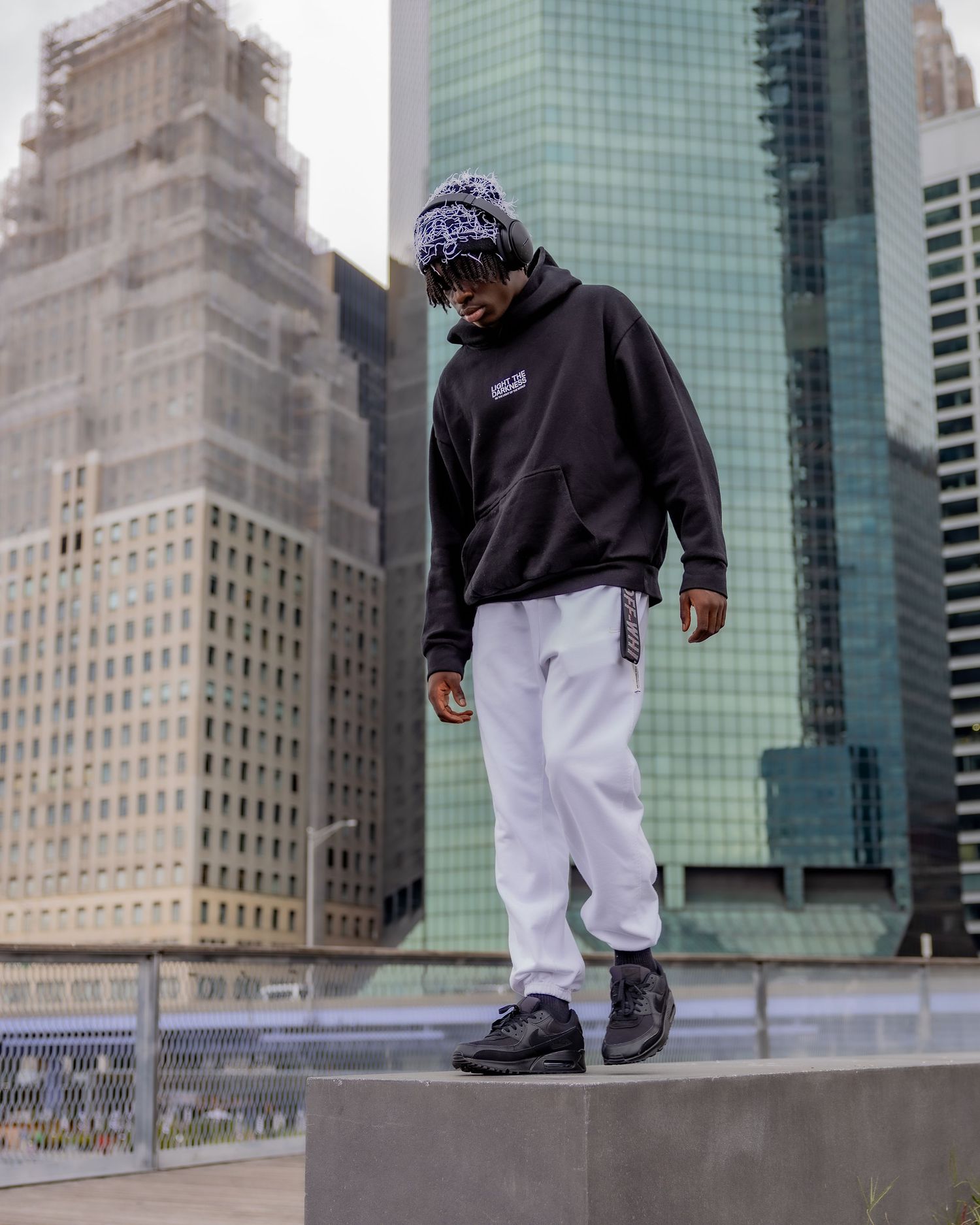 Urban fashion scene with person in black hoodie and white pants against city backdrop.