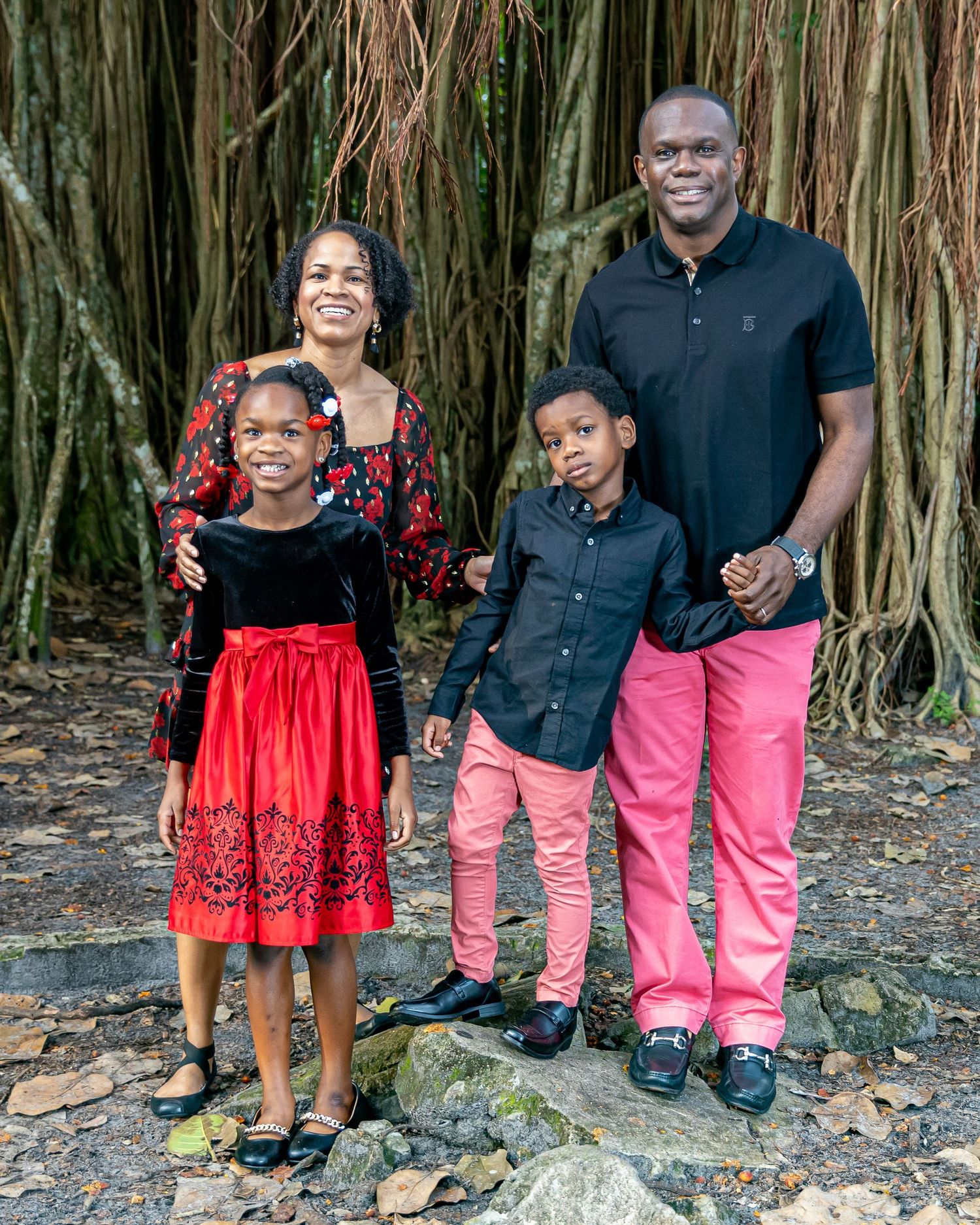 A family poses together against a backdrop of banyan trees wearing coordinated pink and red outfits.
