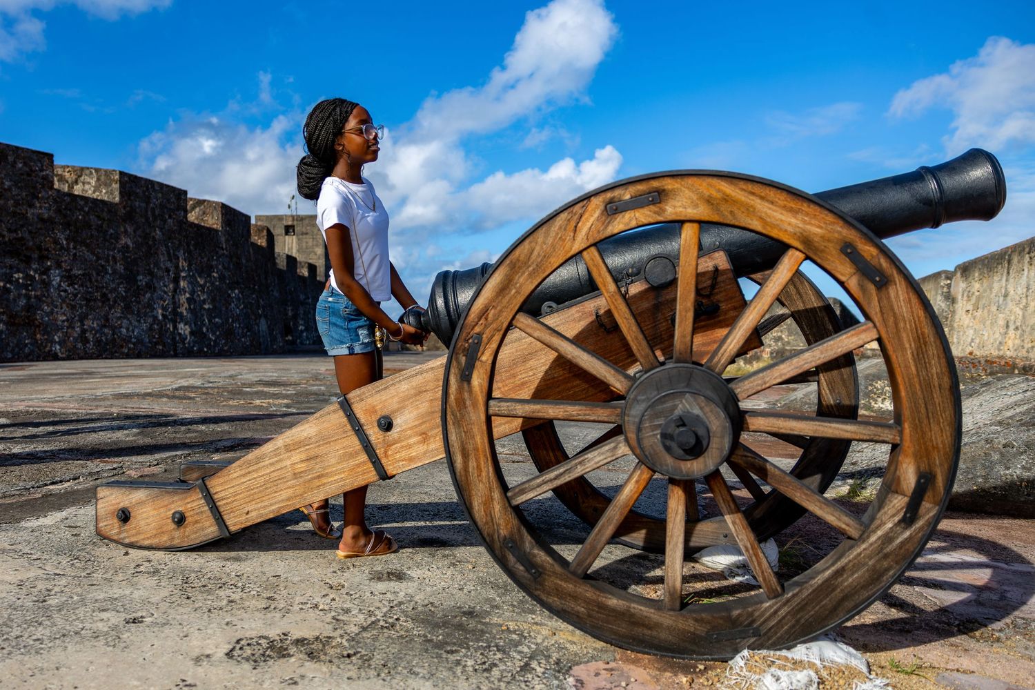 A historic wooden cannon on display at an old stone fortress wall against a blue sky with white clouds.